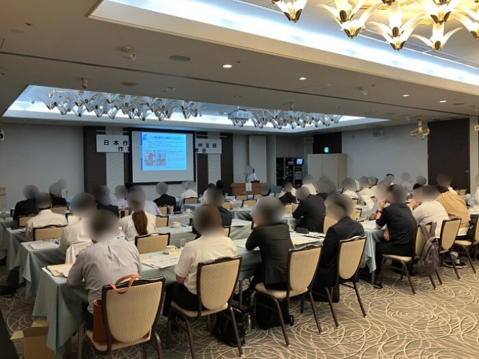 多数の参加者が机に着席して講義を受けている講座会場の後方からの様子。前方にはプロジェクターと講師が立っている。